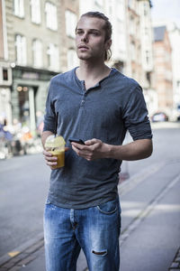 Young man with disposable cup and mobile phone standing on sidewalk