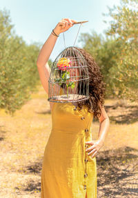 Woman holding birdcage with flower while standing against trees