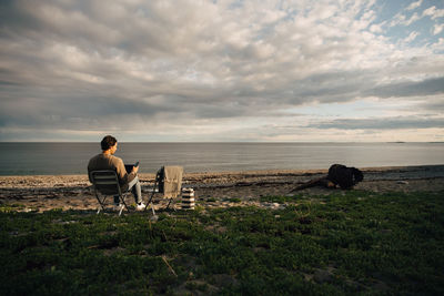 Rear view of woman sitting on chair at sea shore against sky