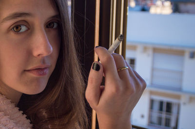 Close-up portrait of young woman holding cigarette by window at home