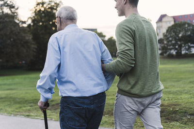 Back view of young man assisting his grandfather walking in a park