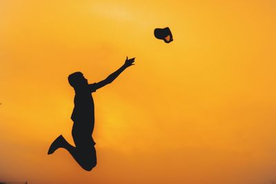 Silhouette man throwing cap while jumping against sky at sunset