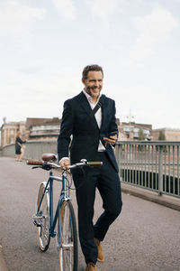 Smiling businessman holding smart phone while walking with bicycle on bridge in city