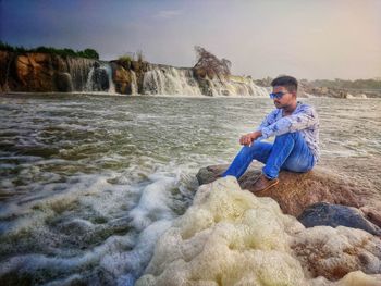 Full length of young man sitting on rock against waterfall