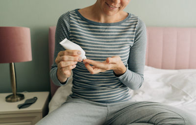 A woman sitting on a bed squeezes cream from a tube onto her finger.