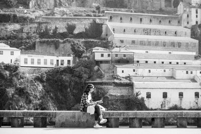 Full length of young woman sitting on retaining wall against buildings