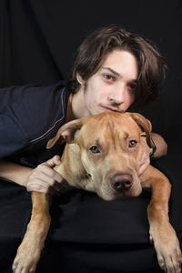 Portrait of a young man with a caramel colored pitbull dog against black background. 