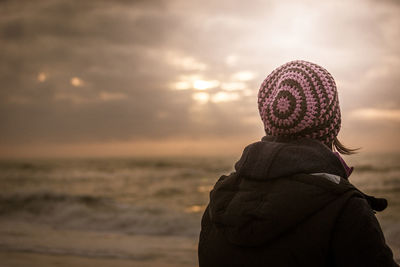 Rear view of woman wearing knit hat at beach against sky during sunset