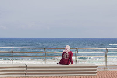 Woman sitting on railing by sea against sky