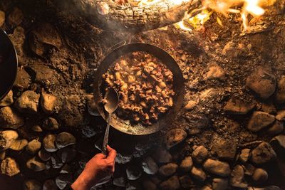 Cropped hand of man cooking food in container by campfire at night