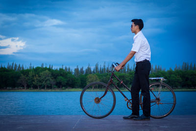 Teenage boy standing with bicycle at lakeshore against sky