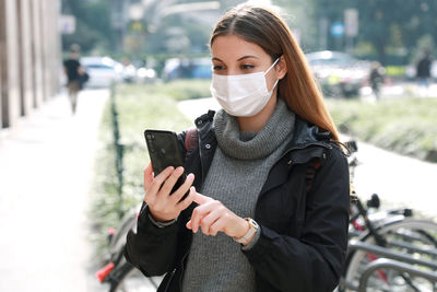 Young woman wearing mask using mobile phone while standing outdoors
