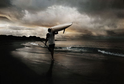 Rear view of man holding kayak walking on beach against sky