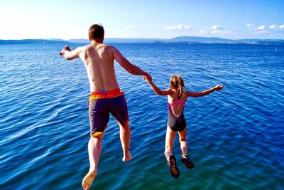 Rear view of shirtless father and daughter jumping in sea against sky