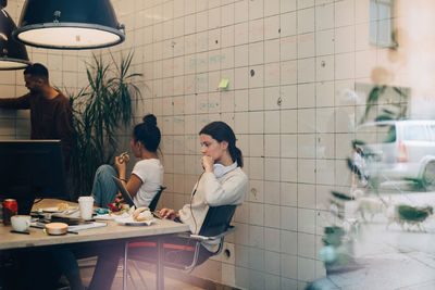 Young businesswoman using laptop while colleagues discussing at creative office seen through window