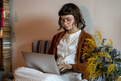 Serious teenage hipster girl sitting comfortably on mattress with laptop working from home