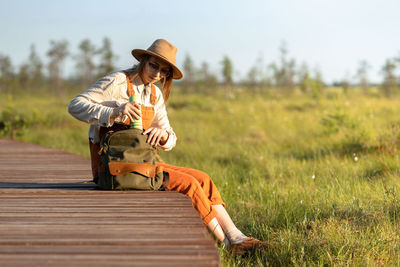 Woman botanist in hat sitting on wooden path through peat bog swamp, takes a thermos from a backpack