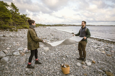 Couple placing blanket on rocky shore at beach during sunset