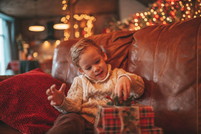 Candid authentic happy child in knitted beige sweater sitting with presents at lodge xmas decorated