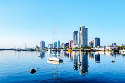 Sailboats in sea against modern buildings in city against clear blue sky