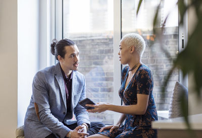 Woman showing mobile phone to colleague while sitting by window in office