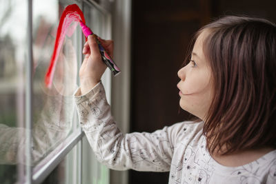 A small serious child draws a rainbow on a window with red marker