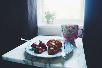 Croissant with strawberries in plate by cup on table