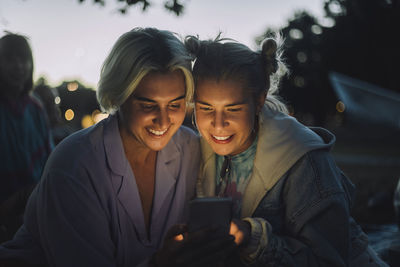 Happy non-binary person sharing smart phone with female friend during sunset