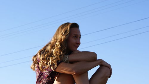 Low angle view of young woman looking away while sitting against clear blue sky during sunny day