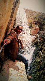 Portrait of man holding monopod while sitting against waterfall