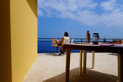 Rear view of woman sitting on chair at building terrace against sky