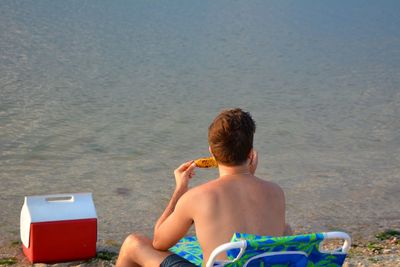 Rear view of shirtless young man eating food while sitting on deck chair at beach