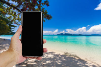 Hand holding mobile phone at beach against sky