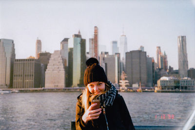 Woman standing by river against modern buildings in city against sky