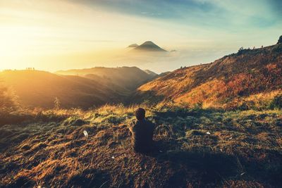 Rear view of silhouette man sitting on mountain against sky