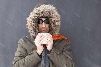 Young man holding coffee cup while standing against wall during snowfall