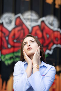 Young woman with hands on chin looking up while standing against graffiti wall
