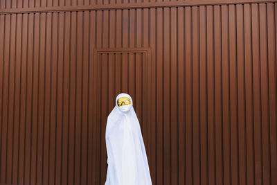 Unrecognizable kid wearing masquerade mask and ghost costume standing in city during halloween and looking at camera