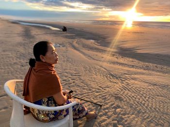 Rear view of girl sitting on sand at beach against sky during sunset