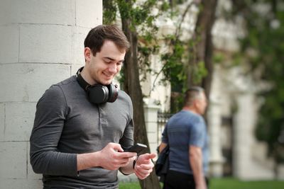 Smiling young man using mobile phone while standing against column