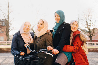 Portrait of happy multi-ethnic female muslim friends with bicycle in city against sky