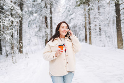 Smiling woman with dark hair in winter clothes using mobile phone in snowy winter forest