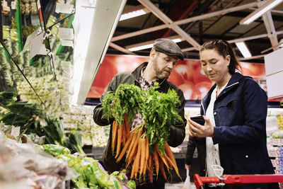 Couple using smart phone while buying carrot at supermarket