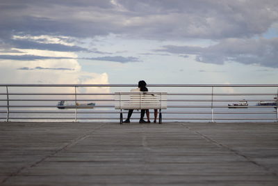 Rear view of couple sitting on bench by railing against sky