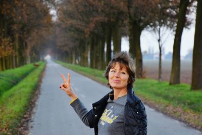 Portrait of mature woman gesturing peace sign on road amidst trees