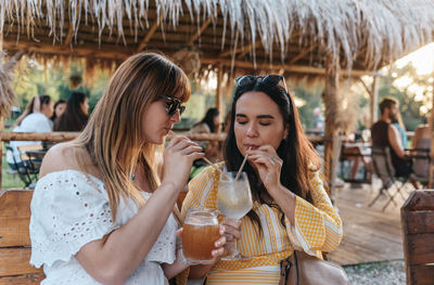 Two women sitting in beach bar at golden hour, drinking cocktails on a straw