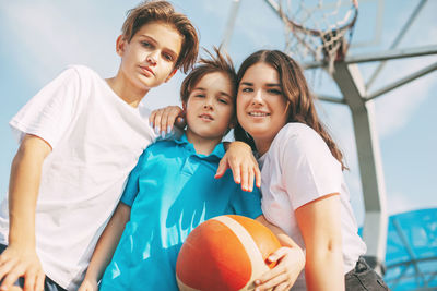 Portrait of smiling siblings with basketball standing against sky