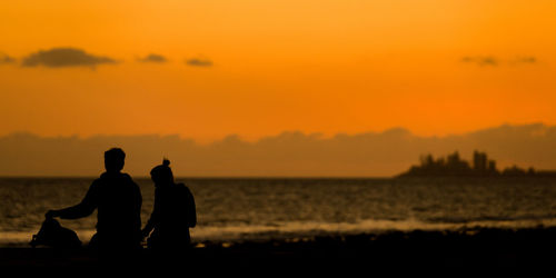 Silhouette couple sitting at beach against orange sky