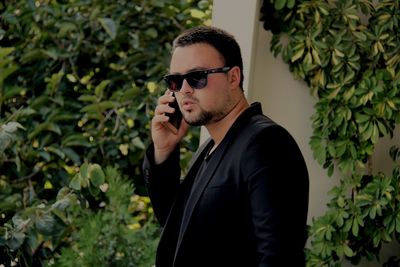 Young man wearing sunglasses talking on smart phone while standing by plants