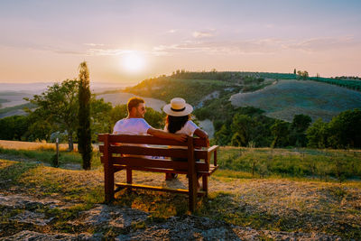 Rear view of couple sitting on bench against sky during sunset